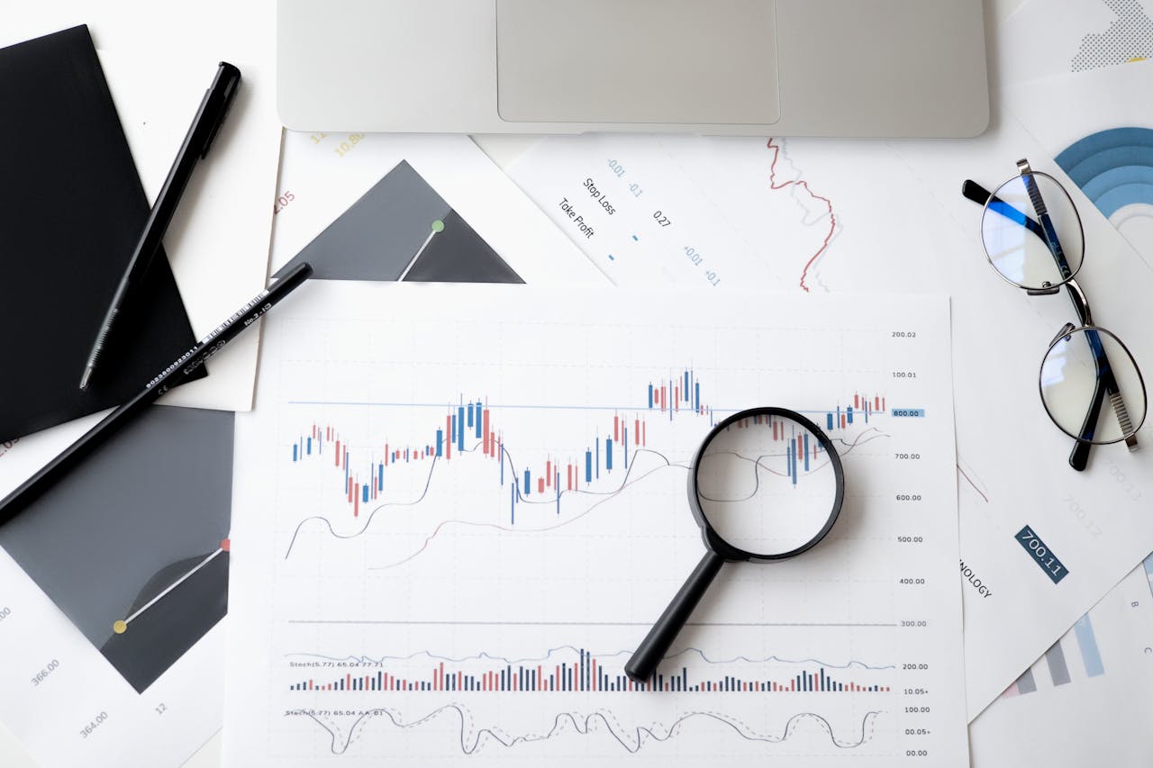 pexels-photo-6801648 Flat lay of stock market analysis documents with magnifying glass, pens, and glasses.